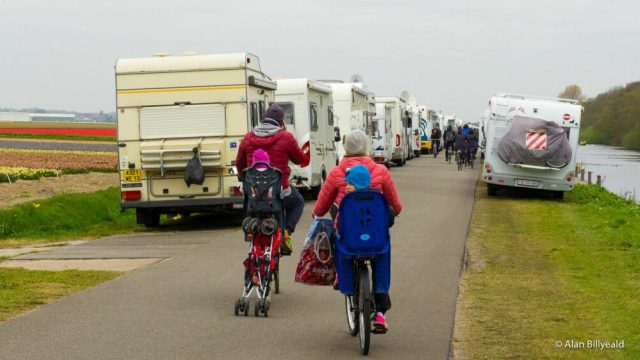 To personer som sykler mellom parkerte bobiler på en smal vei.