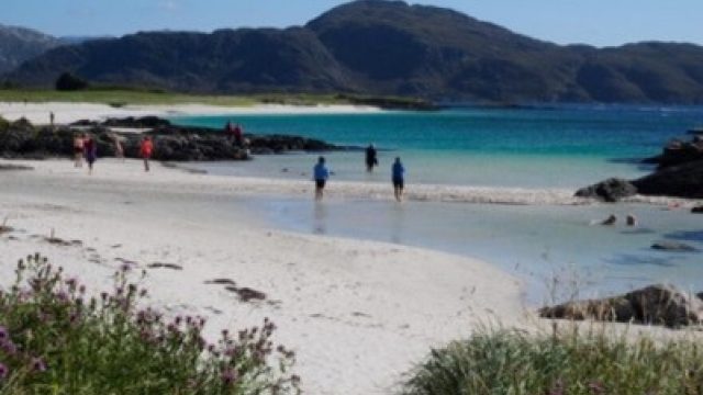 Bilde av personer som vasser i vannkanten på en idyllisk strand med klart, grunt vann og en bakgrunn av majestetiske fjell.