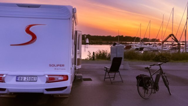 Dette er et bilde som viser en hvit bobil parkert ved siden av en rolig vannkant, sannsynligvis en marina eller en innsjø, ved solnedgang.